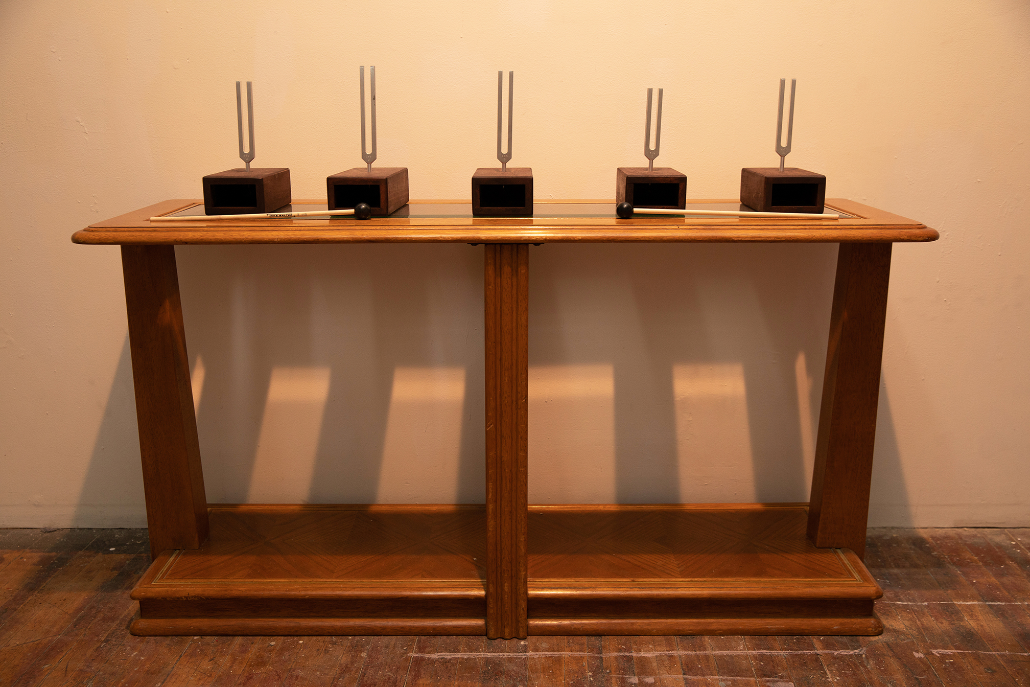 Detail of a small wooden table with five tuning forks set in resanator boxes and two mallets set up on it against a white wall
